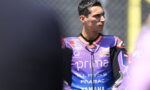 MotoGP rider in a purple Pramac Yamaha racing suit looks to the right, standing by the track fence.