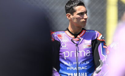 MotoGP rider in a purple Pramac Yamaha racing suit looks to the right, standing by the track fence.