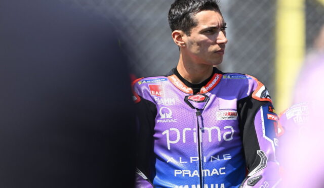 MotoGP rider in a purple Pramac Yamaha racing suit looks to the right, standing by the track fence.