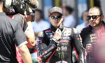 MotoGP rider in black Aprilia racing suit with Monster logo wearing cap and sunglasses, being filmed by a camera crew.