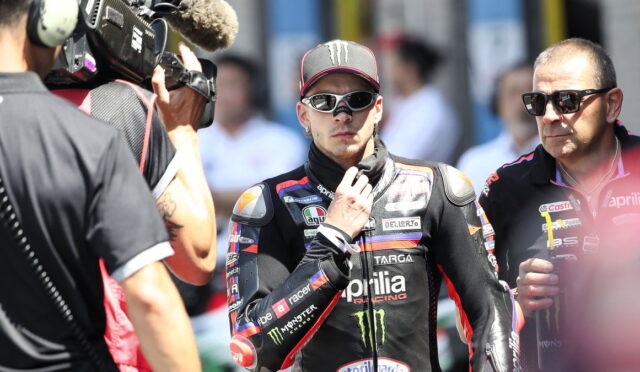MotoGP rider in black Aprilia racing suit with Monster logo wearing cap and sunglasses, being filmed by a camera crew.