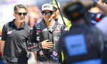 Racing driver in a black suit with logos, wearing sunglasses and a cap, stands beside a man in a black polo shirt during a sunny race event.