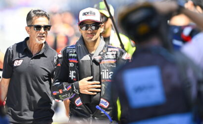 Racing driver in a black suit with logos, wearing sunglasses and a cap, stands beside a man in a black polo shirt during a sunny race event.