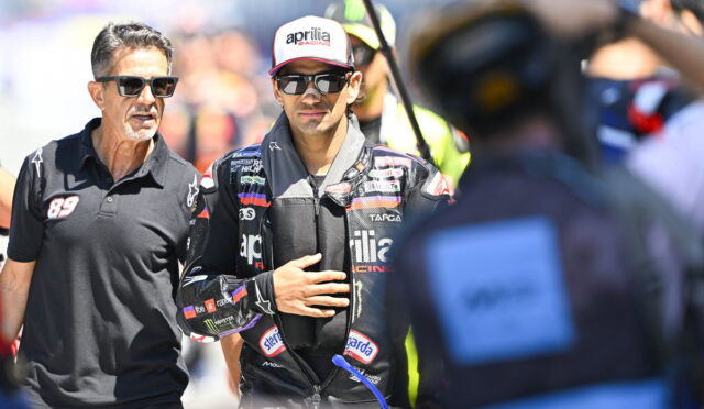 Racing driver in a black suit with logos, wearing sunglasses and a cap, stands beside a man in a black polo shirt during a sunny race event.