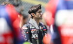 Side view of a male motorcycle racer in a sponsor-logo racing jacket, standing among a blurred crowd.
