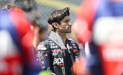 Side view of a male motorcycle racer in a sponsor-logo racing jacket, standing among a blurred crowd.