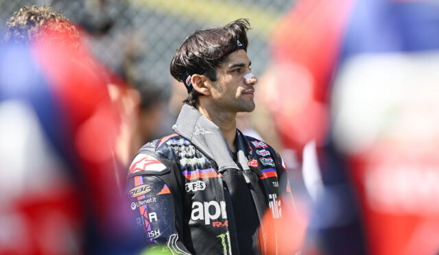 Side view of a male motorcycle racer in a sponsor-logo racing jacket, standing among a blurred crowd.