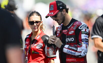 MotoGP rider in red and black racing suit and cap stands with a female team member in a red polo, both looking focused.