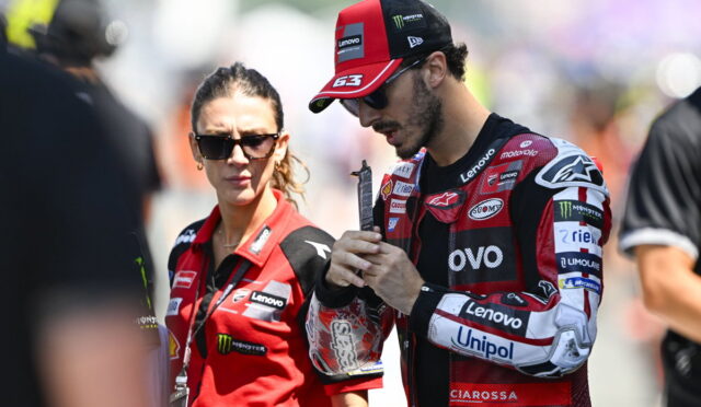 MotoGP rider in red and black racing suit and cap stands with a female team member in a red polo, both looking focused.