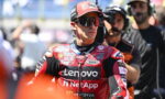 MotoGP rider in a red Lenovo NetApp racing suit and cap, wearing sunglasses, standing in the pit area with crew in the background.