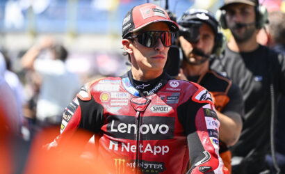 MotoGP rider in a red Lenovo NetApp racing suit and cap, wearing sunglasses, standing in the pit area with crew in the background.