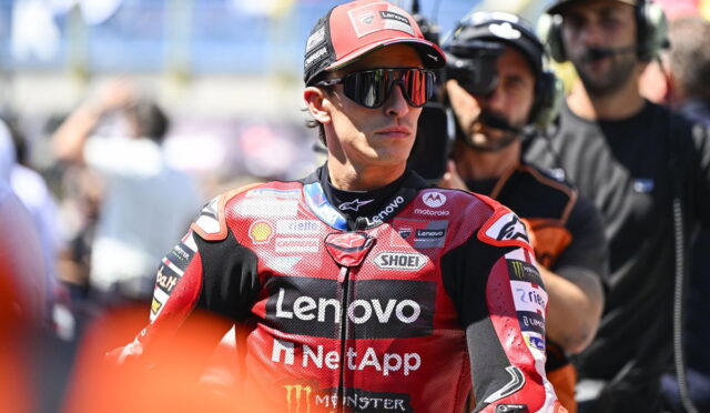 MotoGP rider in a red Lenovo NetApp racing suit and cap, wearing sunglasses, standing in the pit area with crew in the background.