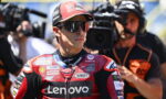 Male motorcycle racer in a red Lenovo racing suit and cap, wearing sunglasses, as cameras film him nearby.