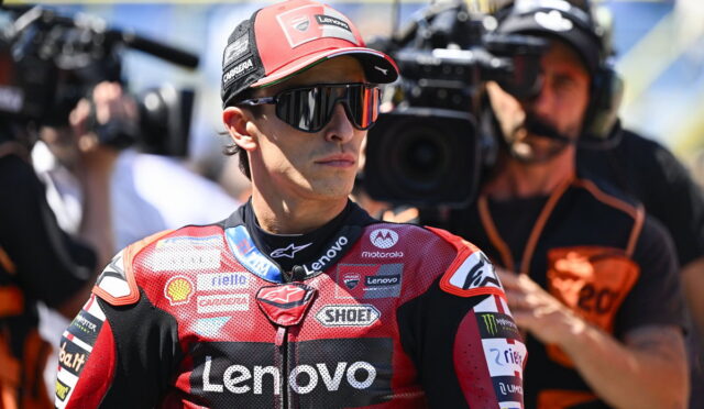 Male motorcycle racer in a red Lenovo racing suit and cap, wearing sunglasses, as cameras film him nearby.