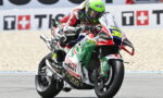 Motocycle racer leaning into a turn on a sport bike with a green helmet and number 36, racing on a track with sponsor decals on the bike and background wall banners.