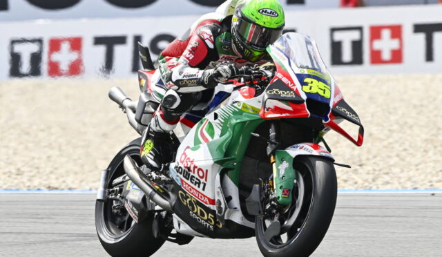 Motocycle racer leaning into a turn on a sport bike with a green helmet and number 36, racing on a track with sponsor decals on the bike and background wall banners.