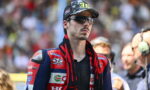Male motorcycle racer in a red-blue racing suit, camo cap and reflective sunglasses at the track, with sponsor patches visible on the suit.
