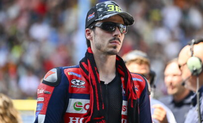 Male motorcycle racer in a red-blue racing suit, camo cap and reflective sunglasses at the track, with sponsor patches visible on the suit.