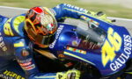 Motorcycle racer in blue and yellow gear leans into a turn on a blue racing bike with sponsor logos such as Gauloises and a prominent number 46 on the fairing with a colorful helmet nearby.