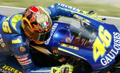 Motorcycle racer in blue and yellow gear leans into a turn on a blue racing bike with sponsor logos such as Gauloises and a prominent number 46 on the fairing with a colorful helmet nearby.