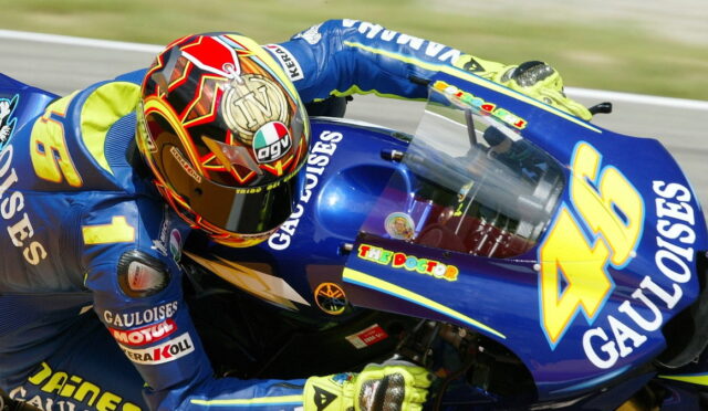 Motorcycle racer in blue and yellow gear leans into a turn on a blue racing bike with sponsor logos such as Gauloises and a prominent number 46 on the fairing with a colorful helmet nearby.