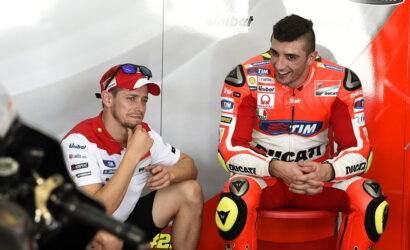 Two motorcycle racers sit in a garage area, one in a white and red polo and cap, the other in red Ducati racing gear on a red stool.