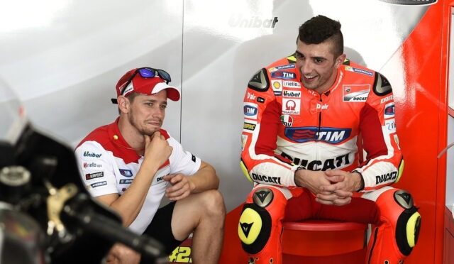 Two motorcycle racers sit in a garage area, one in a white and red polo and cap, the other in red Ducati racing gear on a red stool.