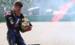 Moto rider in racing suit covers face with hands while holding helmet; red smoke trails behind on a gravel track