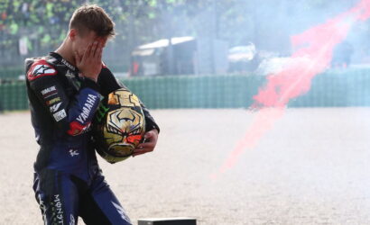 Moto rider in racing suit covers face with hands while holding helmet; red smoke trails behind on a gravel track