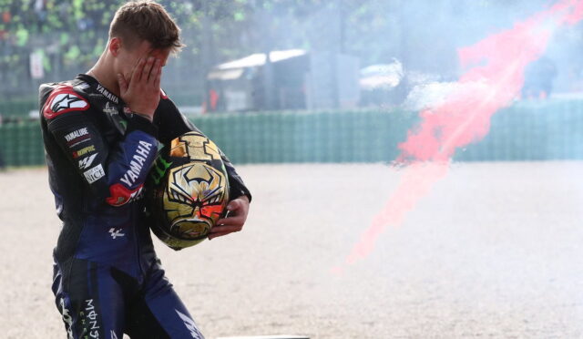 Moto rider in racing suit covers face with hands while holding helmet; red smoke trails behind on a gravel track