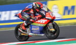 Motorcycle racer in red-blue suit leaning into a turn on a race track with a sponsor-labeled bike in red, blue, and white.
