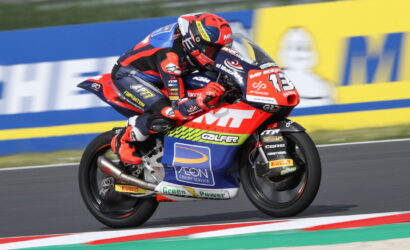 Motorcycle racer in red-blue suit leaning into a turn on a race track with a sponsor-labeled bike in red, blue, and white.