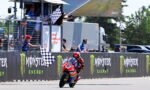 Motorcycle racer wearing red and blue gear crosses the finish line as a checkered flag waves, with Monster Energy banners along the track wall.