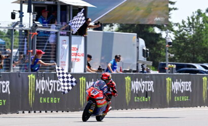 Motorcycle racer wearing red and blue gear crosses the finish line as a checkered flag waves, with Monster Energy banners along the track wall.