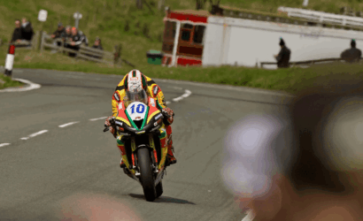 Motorcycle racer wearing a colorful yellow/red suit, number 10, leaning into a road curve with spectators in the background.
