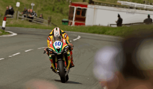 Motorcycle racer wearing a colorful yellow/red suit, number 10, leaning into a road curve with spectators in the background.