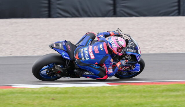 Motorcycle racer in blue and pink suit leaning into a sharp turn on a racetrack with a blue race bike nearby helmet pink visor