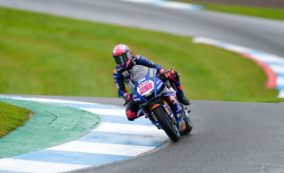Moto racer in blue racing suit on a blue Yamaha bike #28 leaning into a curve on a wet race track, pink helmet visible.