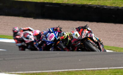 Group of motorcycle racers leaned into a sharp corner on a race track, colorful helmets and suits visible as they hug the inside line.