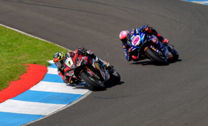 Two motorcycle racers lean into a sharp turn on a racetrack, bikes numbered 7 and 28 close together on a blue-and-white curbed edge.
