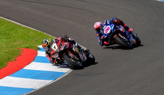 Two motorcycle racers lean into a sharp turn on a racetrack, bikes numbered 7 and 28 close together on a blue-and-white curbed edge.