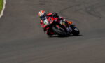 Motorcycle racer in red and black gear leans into a sharp turn on a paved racetrack.