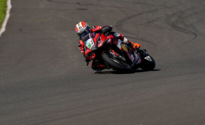 Motorcycle racer in red and black gear leans into a sharp turn on a paved racetrack.