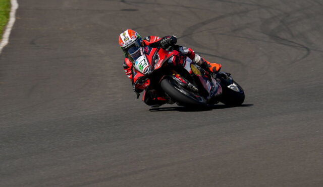 Motorcycle racer in red and black gear leans into a sharp turn on a paved racetrack.