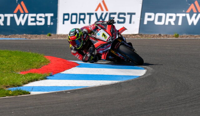 Motorcycle racer in red and black gear leans into a sharp turn on a race track, blue and white curbing visible along the corner.