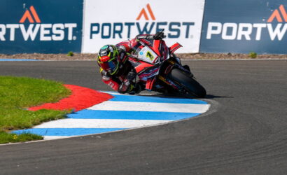MotoGP rider leaning into a sharp turn on a race track, red-white suit and bike, Portwest banners in background