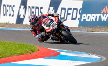 Moto racer in red-and-white gear leans into a high-speed turn on a race track, with blue and white curbs and sponsor boards in the background.