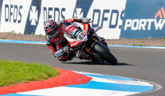Moto racer in red-and-white gear leans into a high-speed turn on a race track, with blue and white curbs and sponsor boards in the background.