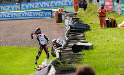 Motocross rider in a red-white racing suit raises an arm in celebration beside a tire barrier on the track, with officials in orange nearby.