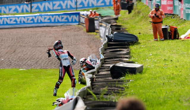 Motocross rider in a red-white racing suit raises an arm in celebration beside a tire barrier on the track, with officials in orange nearby.